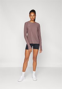 Top deportivo de manga larga en color malva con corte ajustado y escote redondo; combinado con pantalones cortos negros. La modelo lleva zapatillas blancas y calcetines hasta el tobillo.