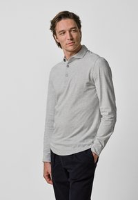 Langärmliges graues Poloshirt aus weichem Stoff, mit einer Knopfleiste mit drei Knöpfen und einem abgerundeten Saum. Getragen mit dunkel gefärbten Hosen.