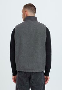 Grau gefütterte Weste mit hohem Kragen, strukturiertem Material und ärmellosem Design. Kombiniert mit einem schwarzen Langarmshirt und dunklen Jeans.