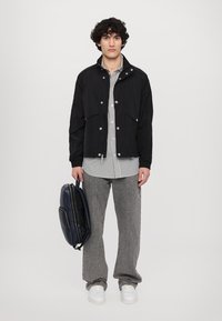 Black lightweight jacket with snap buttons, striped shirt underneath, gray wide-leg pants, and white sneakers. Holding a navy backpack.