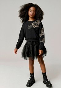 Sudadera negra con bordado de dragón dorado, combinada con una falda de tul negra en capas y botas de combate negras. Pelo rizado.