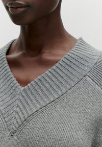 V-Ausschnitt Pullover in Hellgrau, mit geripptem Kragen und strukturiertem Stoff. Eine Nahaufnahme hebt die Nähte und Details des Halsausschnitts hervor.