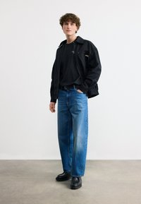 Νεαρός άνδρας με σγουρά μαλλιά, φοράει μαύρο μπουφάν, μαύρο πουκάμισο, μπλε τζιν και μαύρα παπούτσια, στέκεται μπροστά σε έναν απλό άσπρο τοίχο.