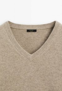 Pull en laine beige à col en V avec encolure côtelée et petite étiquette de marque noire à l'intérieur du col.