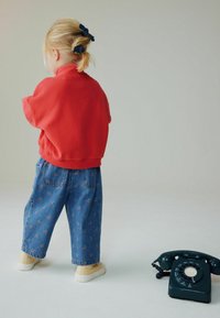 Sudadera roja, jeans de mezclilla con patrón floral y zapatos beige. Un teléfono negro vintage de disco está colocado cerca sobre un fondo claro.