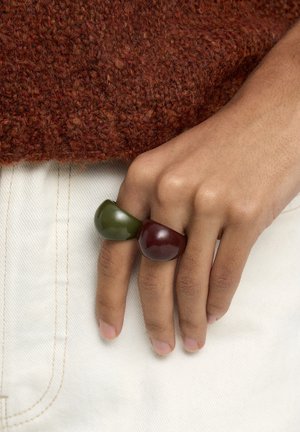 Ręka nosząca dwa masywne pierścionki w kolorze oliwkowej zieleni i głębokiej czerwieni, spoczywająca na białych spodniach z teksturowanym, rdzawej barwy swetrem.