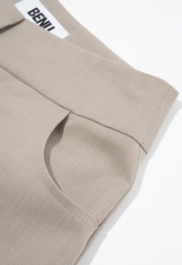 Beige broeken met een gladde stoftextuur, voorzien van een omgevouwen tailleband en zijzakken. Etiket met de tekst "BENILI" aan de bovenkant.