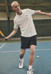 Λευκό πόλο μπλουζάκι με γιακά και μαύρο λογότυπο, συνδυασμένο με μαύρες βερμούδες. Παίκτης σε κίνηση σε γήπεδο τένις με ρακέτα και μπάλα τένις.