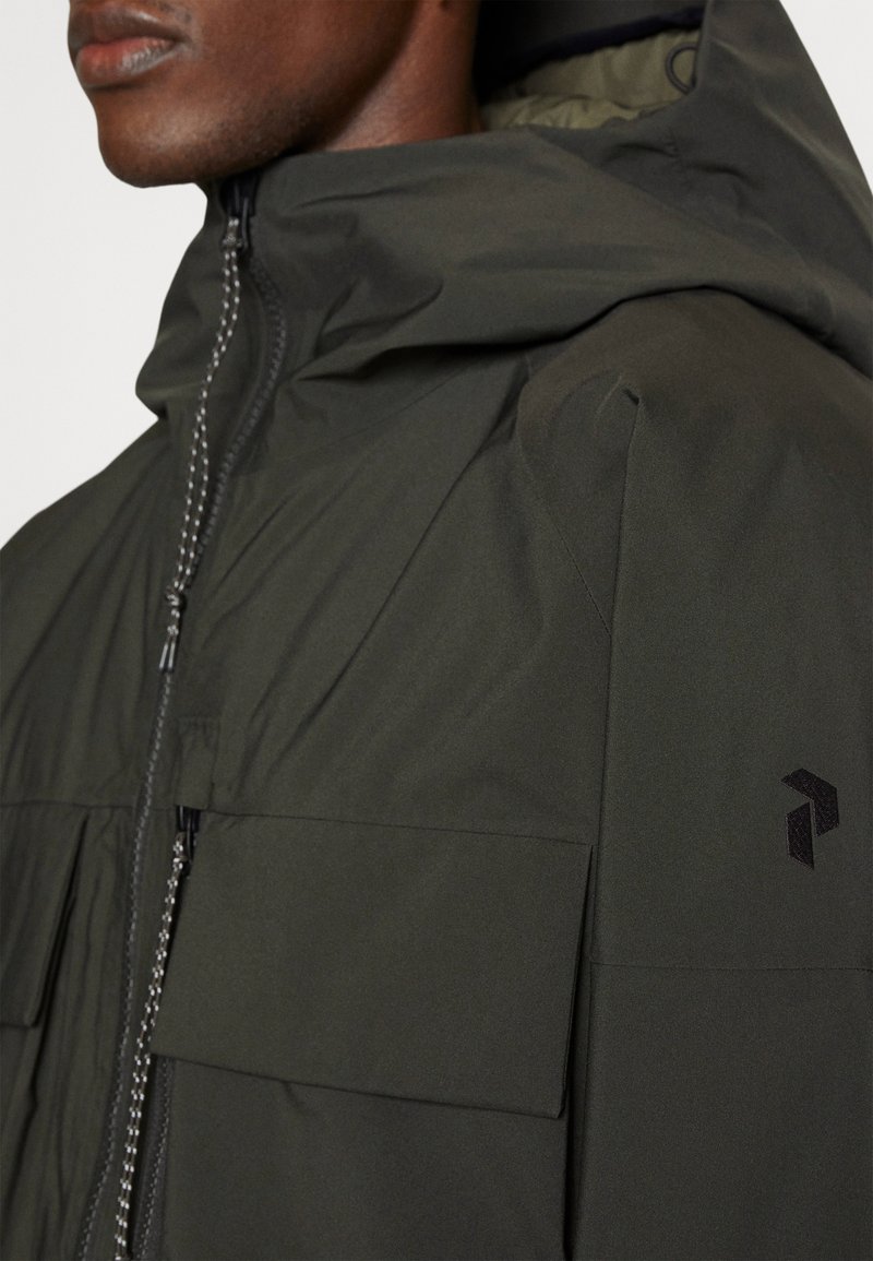 Chaqueta verde oscuro con cierre de cremallera, grandes bolsillos frontales y capucha. Presenta un logotipo texturizado en la manga y un acabado de tela suave.