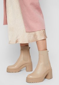 Botines de cuero beige con paneles laterales elásticos y tacón grueso de bloque. La suela presenta un patrón de dibujo resistente.