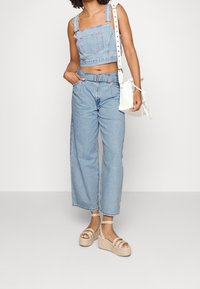 Džínová crop halenka s nastavitelnými ramínky, světle modré široké kalhoty a béžové platformové espadrilky. Doplňky zahrnují bílou tašku.