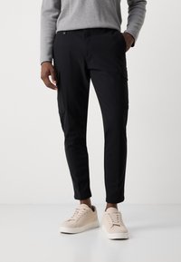 Zwarte cargobroek met een slim fit, voorzien van zakken aan de zijkanten, gemaakt van lichtgewicht stof. Gecombineerd met beige suède sneakers.