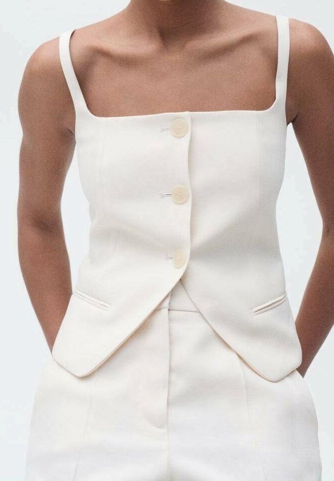 Femme portant un gilet blanc sans manches sur mesure avec trois boutons et un pantalon blanc taille haute assorti, mains dans les poches.