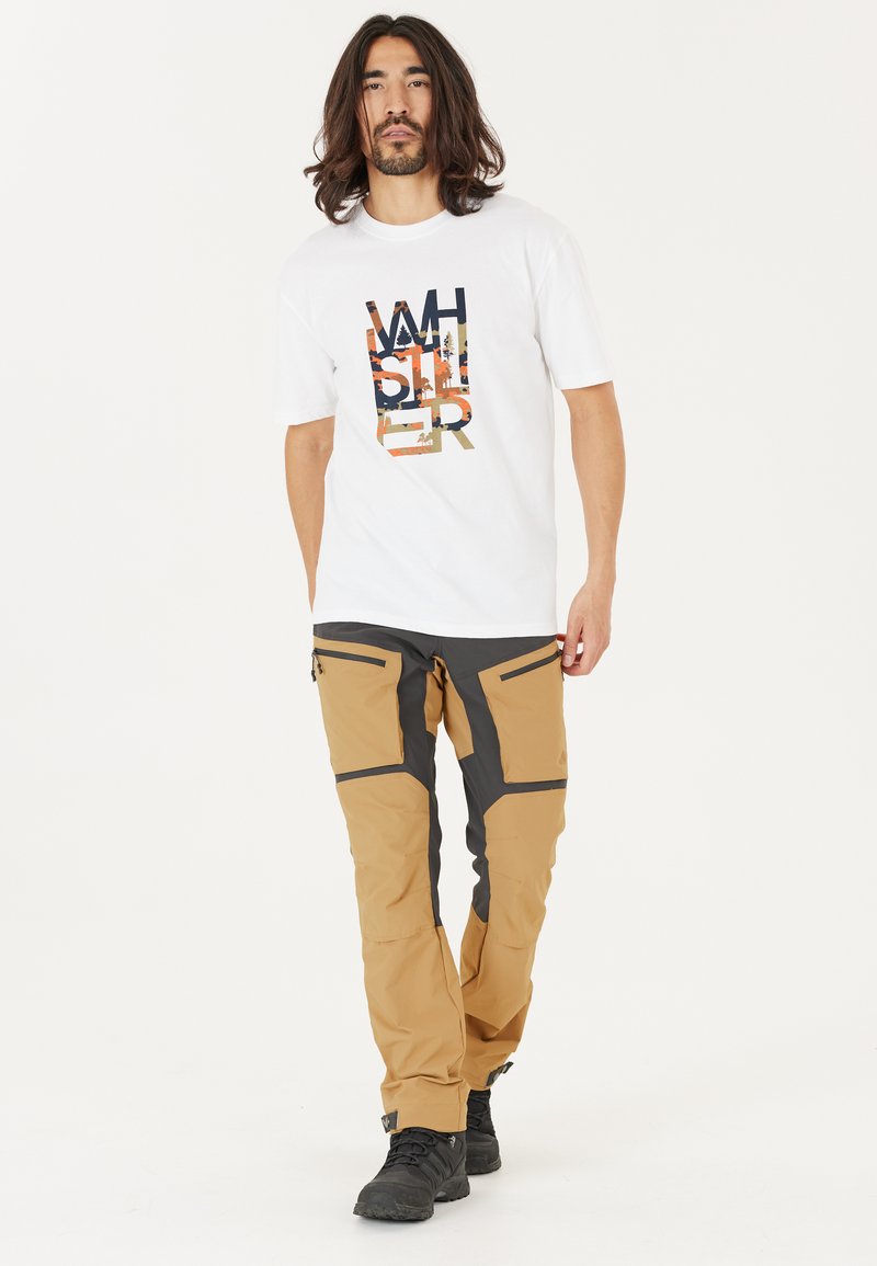 Witte grafische T-shirt met kleurrijk "Whistler"-ontwerp; tan en grijze cargobroek met meerdere ritszakken en een gestructureerde stof. Zwarte laarzen.