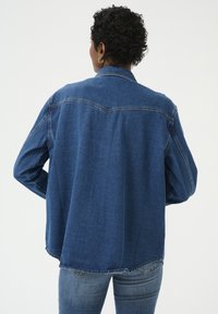 Tumši zilas džinsa jaka ar klasisku apkakli, garām piedurknēm un nedaudz izliekta apakšmali. Mugurpusē redzamas šuvju detaļas un brīvi pieguloša forma.