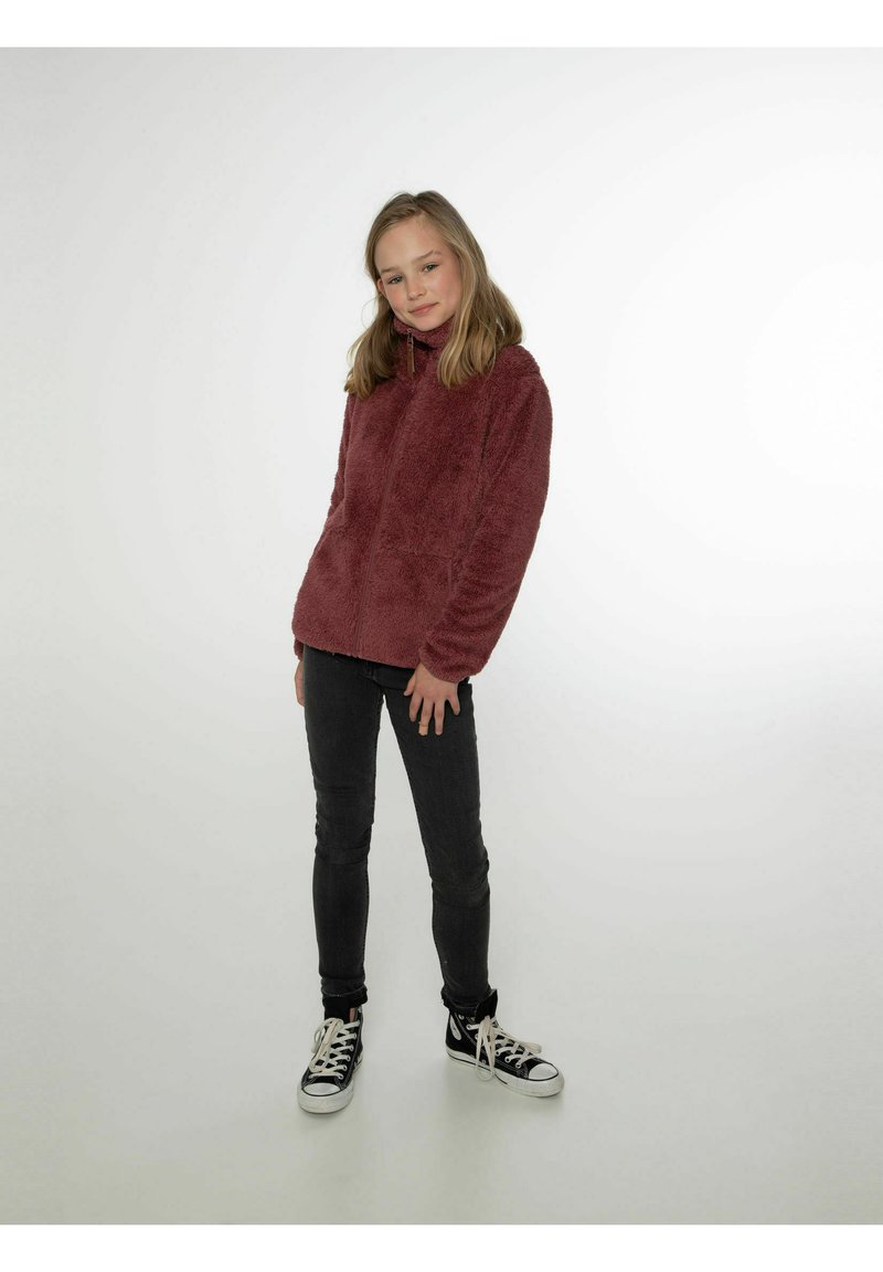Giacca in pile bordeaux con colletto alto e zip frontale, dotata di due grandi tasche. Indossata con jeans skinny neri e sneaker nere.