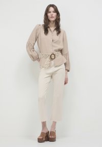 Camicia button-up di un marrone chiaro con maniche a sbuffo e una cintura intrecciata beige con una fibbia circolare, abbinata a pantaloni cropped crema e scarpe con zeppa marroni.