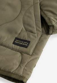 Olivgrön quiltad textil med en mjuk och slät yta. Har kontrasterande sömmar och en etikett med koordinater och varumärke.