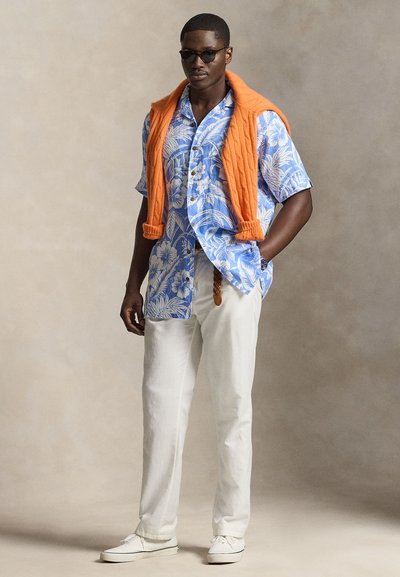 Moški z rjavimi sončnimi očali, modro kratko srajco z rožnatim vzorcem, belimi hlačami, belimi supergami in oranžnim puloverjem, premetanim čez ramena.