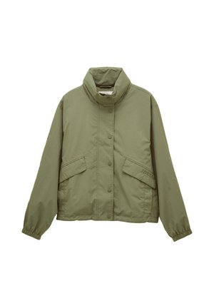 Femme aux cheveux bouclés portant une veste verte, une chemise rose rayée, un jean bleu à revers larges et des baskets orange, debout contre un mur beige.