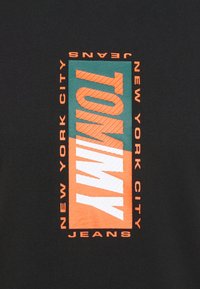 Mustat t-paidat, joissa on rohkea, keskeinen grafiikka, jossa lukee "TOMMY" valkoisella ja oranssilla turkoosilla taustalla, ja "NEW YORK CITY JEANS" oranssilla sivuilla.