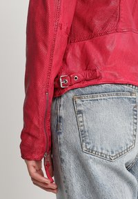 Czerwona skórzana kurtka z teksturowanymi panelami z siateczki, z zamkiem błyskawicznym na rękawie i paskiem z klamrą w talii. Noszona z jasnoniebieskimi dżinsami.