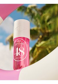 Różowa butelka mgiełki perfum Sol de Janeiro z białą nakrętką, otoczona różowo-białą dmuchaną pływającą podkładką, rozmazane palmy w tle.