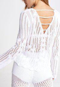 Haut blanc en maille à manches longues, dos ouvert avec des lanières horizontales, et un ourlet festonné. Le tissu présente un tissage texturé et lâche.