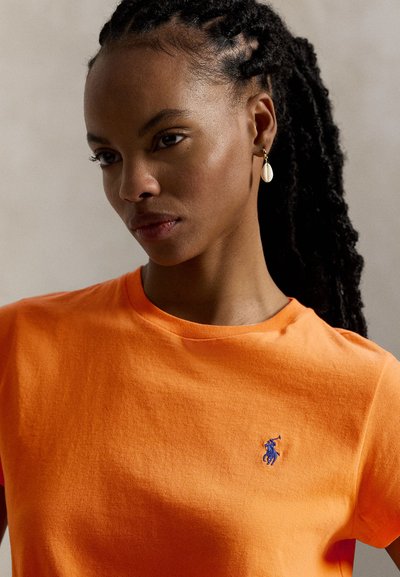 Φωτεινό πορτοκαλί βαμβακερό μπλουζάκι με στρογγυλή λαιμόκοψη, με μπλε κεντητό λογότυπο στο αριστερό στήθος και λεία, αδιάφορη υφή.