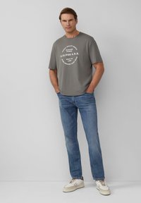 Grijze katoenen T-shirt met witte ronde grafische tekst, gecombineerd met lichtblauwe denim jeans en beige sneakers. Eenvoudige, casual outfit.