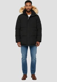 Sort puffervest med hætte foret med pels, med lynlåslukning, store forlommer og quiltet tekstur. Båret over lyseblå jeans og brune sko.