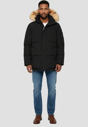 Sort puffervest med hætte foret med pels, med lynlåslukning, store forlommer og quiltet tekstur. Båret over lyseblå jeans og brune sko.