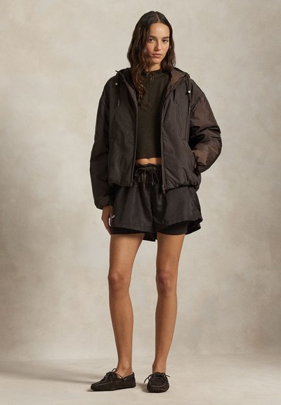 Μπουφάν τύπου puffer μαύρου χρώματος με κουκούλα, ελαφρώς κομμένο τοπ ελιάς, μαύρα σορτς και σκούρα loafers. Απαλές υφές, άνετη εφαρμογή και ρυθμιζόμενη μέση.