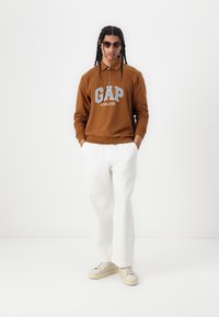Brun huvtröja med "GAP ATHLETIC" logotyp, vita löst sittande byxor och krämfärgade sneakers. Modellen bär solglasögon och har flätat hår.