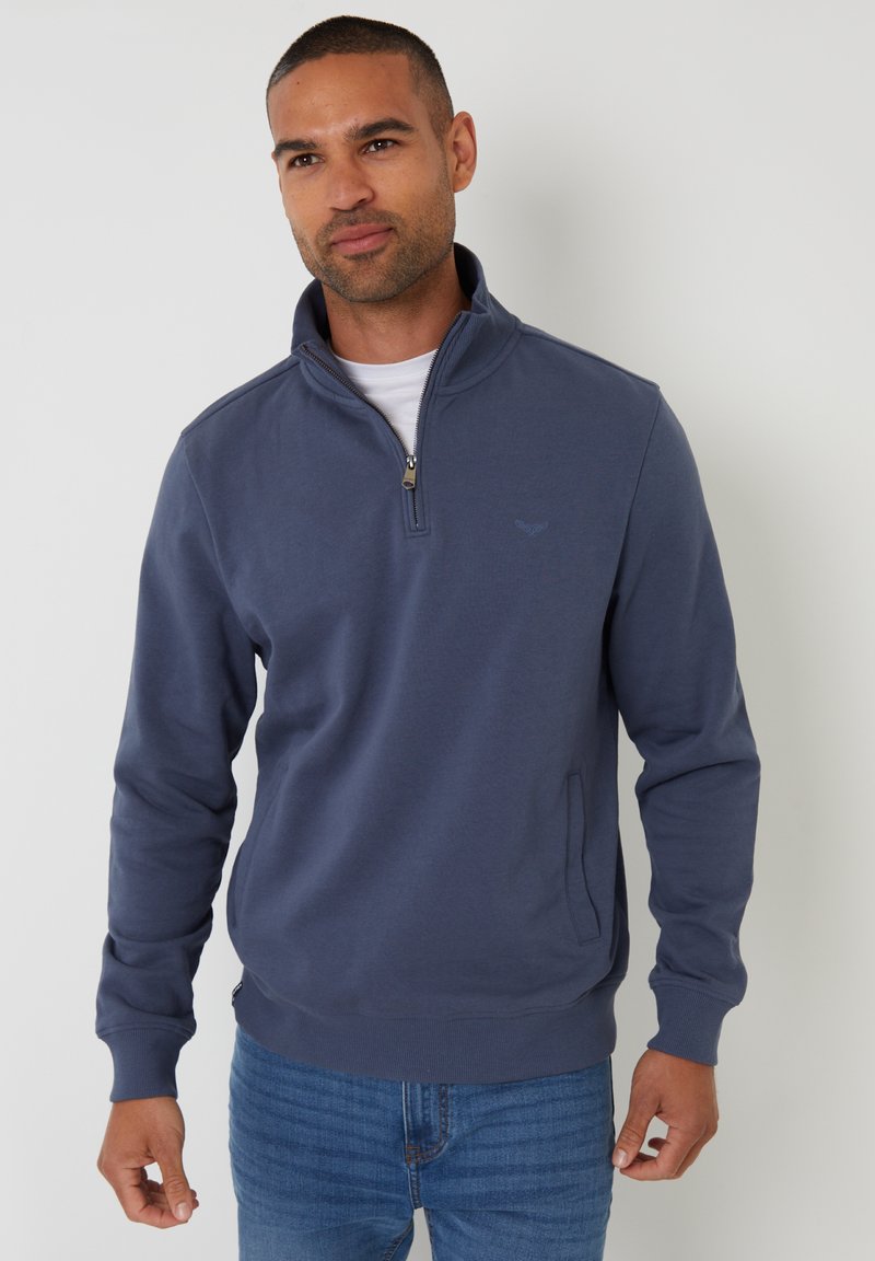 Man draagt een blauw kwart-rits sweatshirt over een wit shirt en een blauwe spijkerbroek, staand tegen een effen lichte achtergrond.