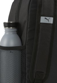 Mochila negra con un bolsillo lateral de malla que sostiene una botella de agua gris. Incluye un parche del logo Puma gris en el lateral.