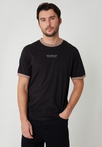 Hombre con camiseta negra con ribete beige y texto "Santa Monica Ride", de pie con una mano en el bolsillo contra un fondo liso.