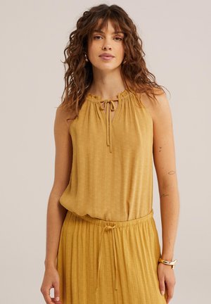 Młoda kobieta o falujących brązowych włosach, ubrana w żółtą sukienkę bez rękawów z zaakcentowaną talią i wiązaniami, stojąca na tle jednolitego beżowego tła.