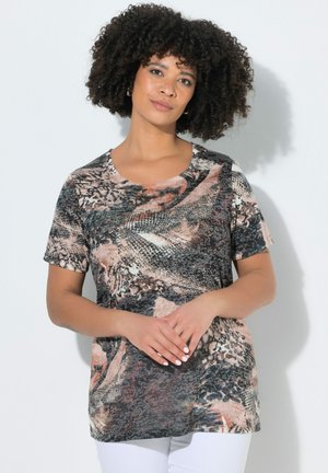 Kvinde med krøllet hår iført en kortærmet, flerfarvet dyreprint-top og hvide bukser, stående foran en ensfarvet lys baggrund.