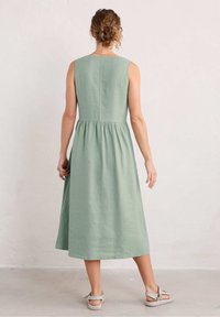 Femme vue de dos, portant une robe midi sans manches de couleur vert clair et des sandales beiges, se tenant contre un mur clair.