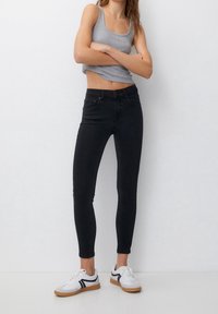 Jeans pitillo negros hechos de mezclilla, con cintura de talle medio y estilo clásico de cinco bolsillos, combinados con una camiseta sin mangas gris y zapatillas blancas.