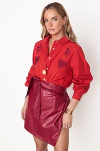 Punainen puuvillapaita, jossa on brodeerattuja sydämiä ja kukkia, yhdistettynä kiiltävään viininpunaisesta keinonahkasta valmistettuun kääntöhameeseen, jossa on sidottu vyötärö.