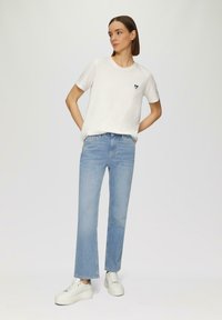 T-shirt blanche à manches courtes avec un emblème en forme de cœur bleu marine, assortie à un jean droit bleu clair et des baskets blanches à semelles texturées.