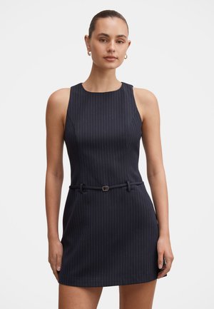 Femme portant une robe noire sans manches à fines rayures, avec un col montant et une fine ceinture assortie à la taille, debout devant un fond uni.