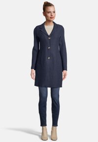 Marineblauer Wollmantel mit Reverskragen, fünf Knöpfen und Seitentaschen, kombiniert mit dunklen Jeans und beigen Stiefeletten.