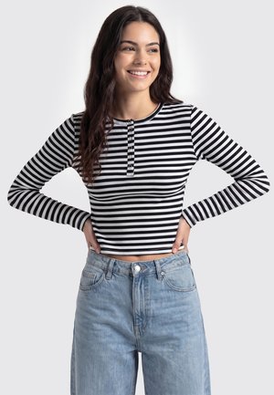 Młoda kobieta uśmiechająca się, z długimi ciemnymi włosami, ubrana w czarno-biały, paskowany, długi crop top z długim rękawem oraz jasnoniebieskie, wysokie jeansy.