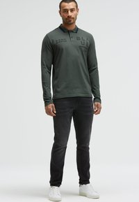 Polo a maniche lunghe verde scuro con colletto blu navy, caratterizzato da dettagli testuali neri. Abbinato a jeans scuri slim-fit e sneaker bianche.