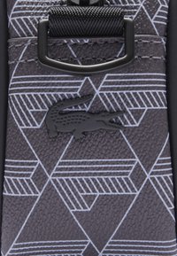 Pattern geometrico nero e grigio su materiale texturizzato con un logo di coccodrillo nero e un dettaglio di fibbia nera.