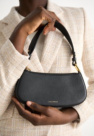 Black leather handbag with a textured finish, sleek design, short strap, and a gold logo accent. Held against a light-colored fabric backdrop.