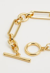 Bracciale in oro con maglie grandi e piccole intrecciate, un’etichetta circolare e una barra orizzontale con dettagli del marchio. Finitura liscia e lucida.